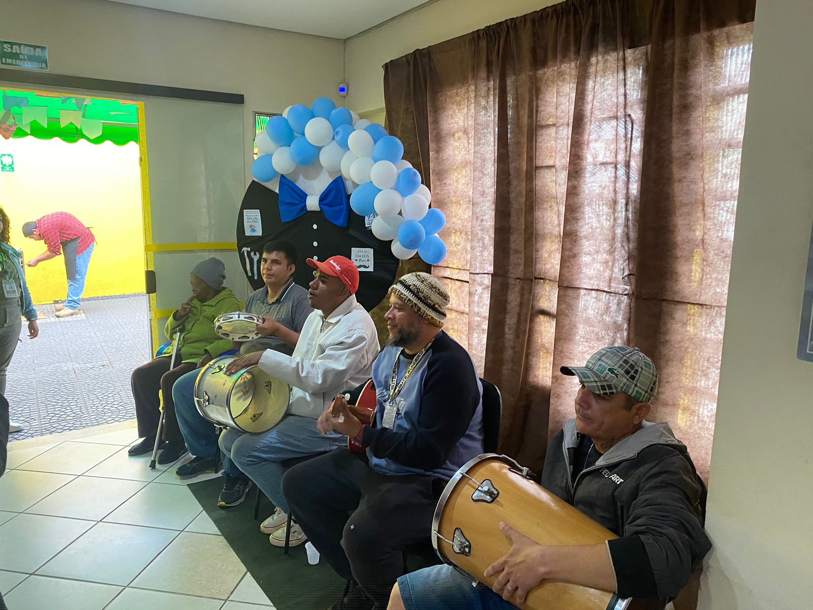 Na imagem, o grupo de pagode que tocou no local, tocando pandeiro, tambor e cavaquinho, na frente de uma cortina marrom e bexigas azuis.